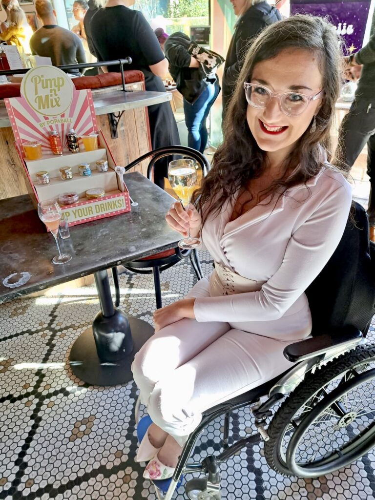 Georgina is a white female in a manual wheelchair. She is holding a glass of prosecco.