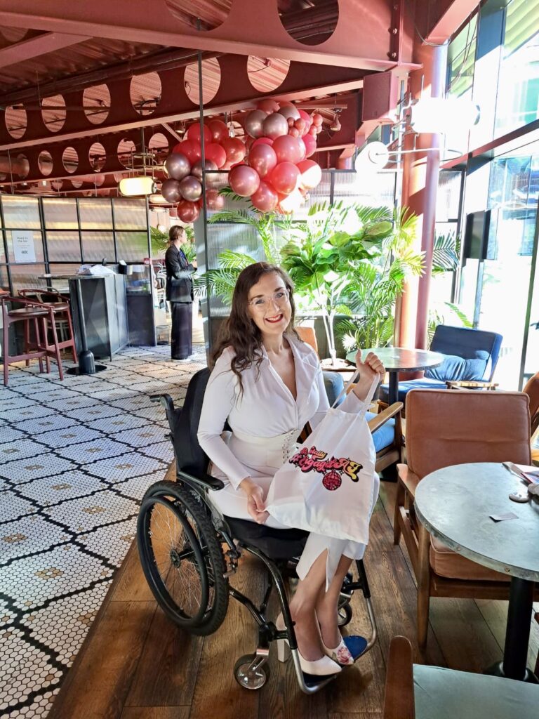 Georgina is a white female in a manual wheelchair. She is holding a Quirky Wedding Fayre goody bag.