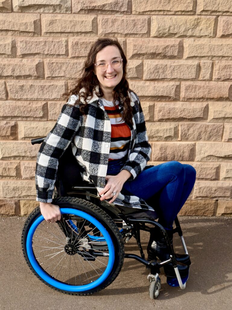 Georgina (a white brunette female) is seated in a wheelchair with blue handrail covers