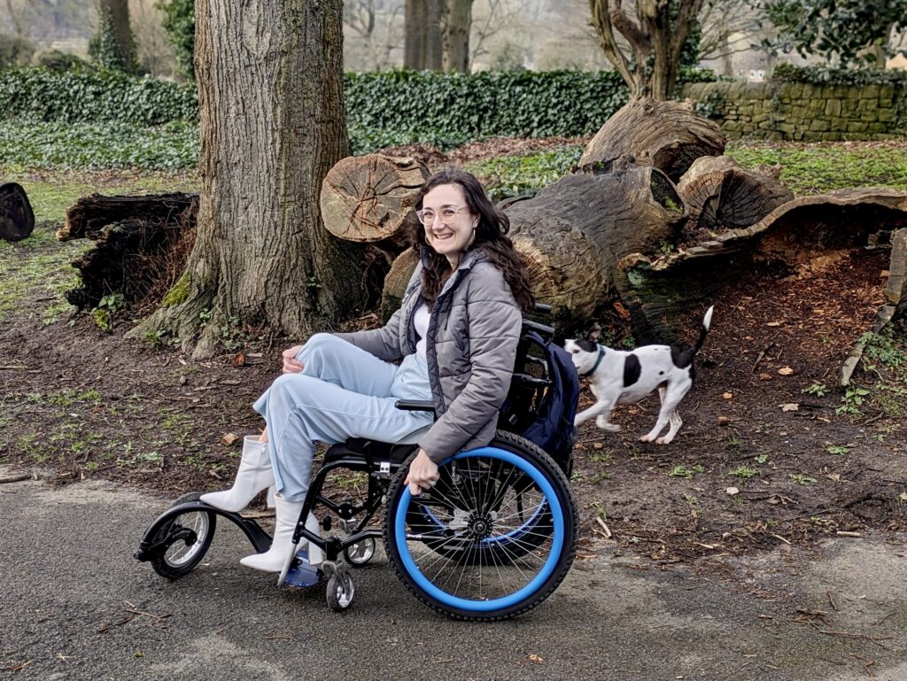 Georgina (a white brunette female) is out walking her black and white staffy dog in her manual wheelchair.