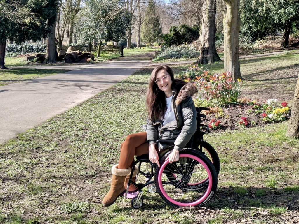 Georgina (a white brunette female) is wearing a green coat and is seated in a manual wheelchair with pink pushrim covers