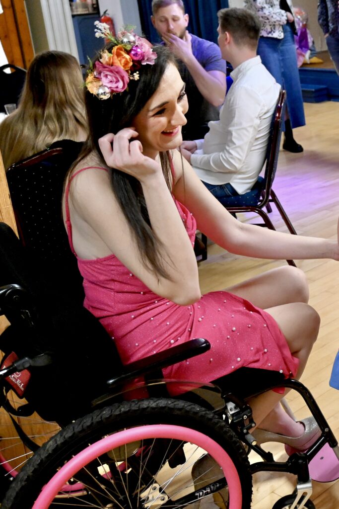 Georgina (a white brunette female) is wearing a pink party dress and has pink handrail covers on her wheelchair