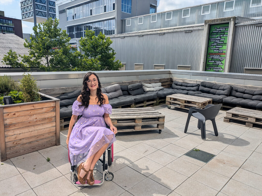 Georgina (white, brunette female in a manual wheelchair) wears a purple dress and is seated in front of a cosy balcony area.