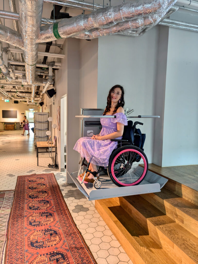 Georgina, white brunette in a manual wheelchair, is in a lilac dress. She is in her wheelchair on the hostel's hoist ramp