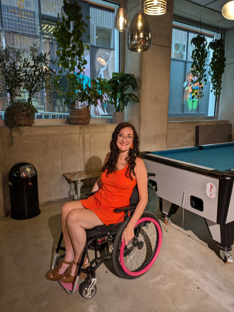 Georgina (white female and brunette) is sat in her manual wheelchair with pink push rims. Behind her is a large pool table