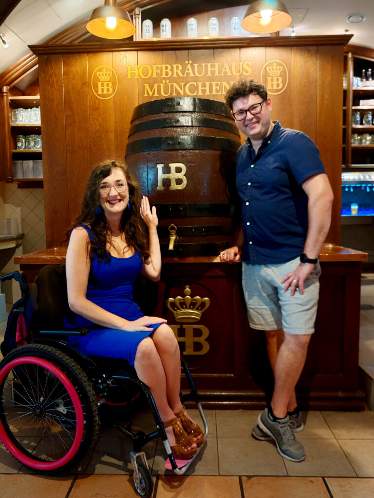 Georgina is seated in her wheelchair with her husband Richard standing beside her. They stand by a large Hofbräuhaus keg