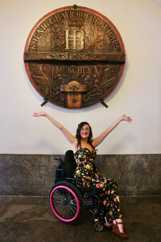 Georgina is a white, female wheelchair user. She has her hands up in front of the Hofbräuhaus beer keg mounted on the wall of their Munich entrance