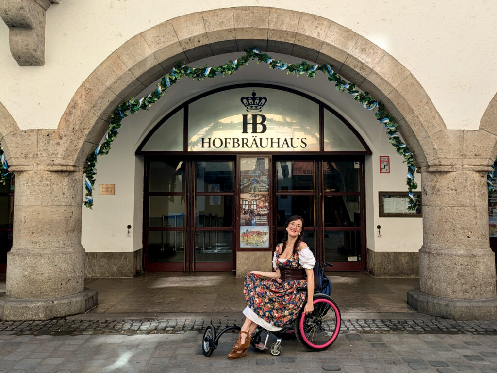 Georgina wears a traditional dirndl outfit and is outside the entrance in Munich in her pink push rimmed wheelchair