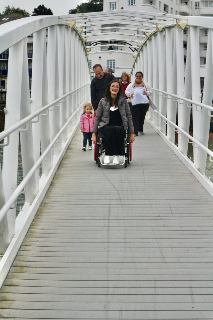 Georgina, a white brunette female, wheels in her manual wheelchair down a bridge ramp over the water in Cowes harbour