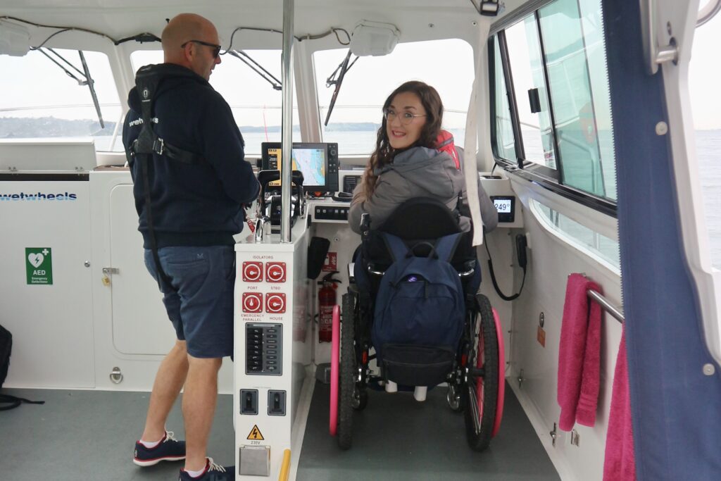 Rear view of Georgina who is seated in her manual wheelchair with pink push rims. She is smiling at the camera as she captains/ sails the wheelchair friendly sailing experience with Wetwheels