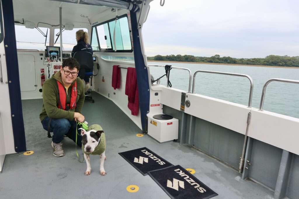 Richard, a white male with brunette hair, smiles and crouches beside Milo who is a black & white staffy dog. They are on a Wetwheels accessible boat.