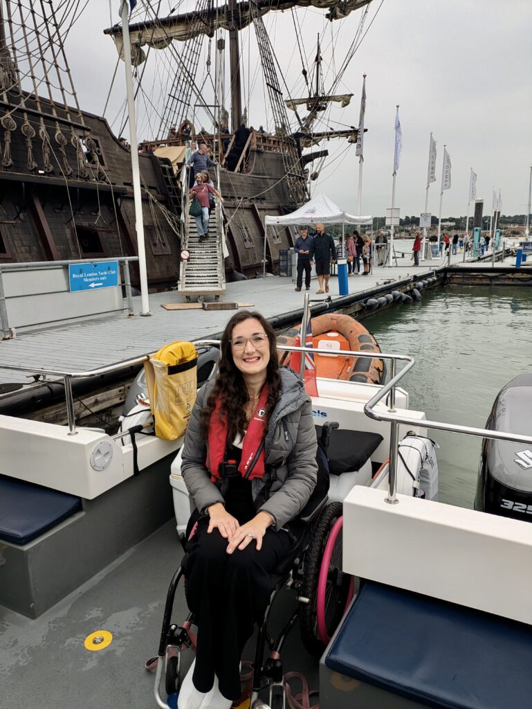 Georgina is seated in a manual wheelchair and is on an adapted sailing boat with Wetwheels. You can see the spanish galleon ship behind her