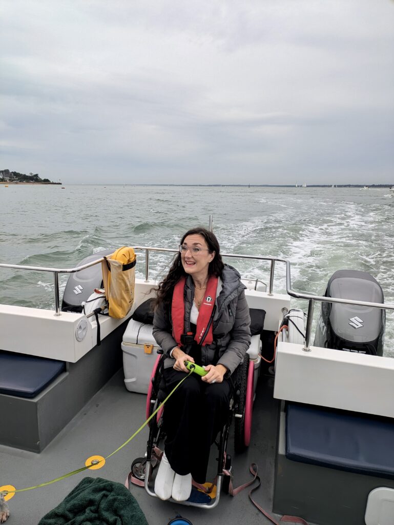 Georgina, a white brunette female, is seated in her wheelchair. This is a candid photos of her with the sea behind her