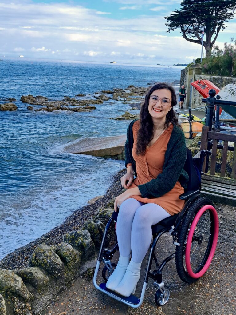 Georgina, a white brunette female who uses a wheelchair, is in a wheelchair accessible location on the Isle of Wight. She is by the beach in the Seaview town and smiles at the camera whilst wearing an orange dress