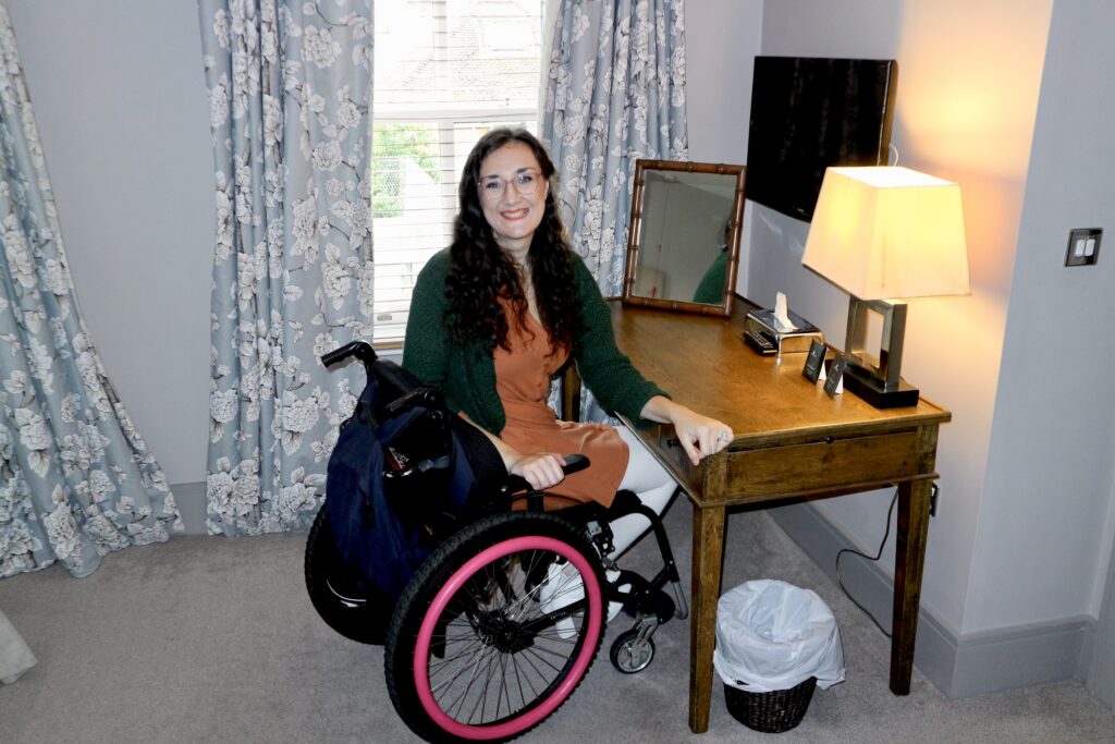 Georgina, a white brunette female, who is seated in a manual wheelchair pulled into a desk area