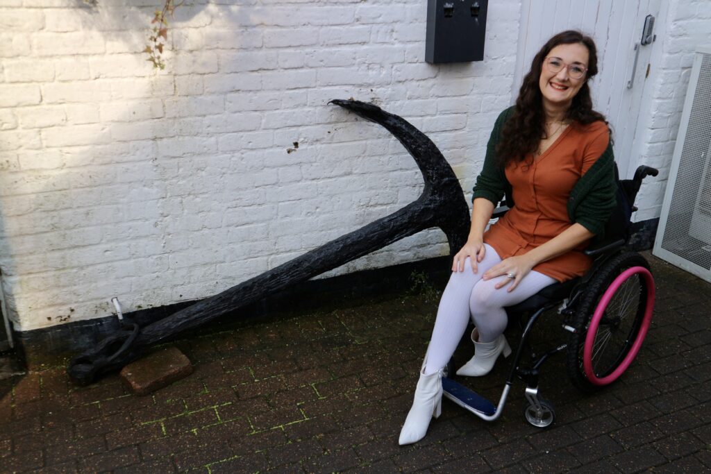 Georgina, a white brunette female who uses a wheelchair, is in the wheelchair accessible Seaview Hotel. She is pictured in front of a retired ships anchor