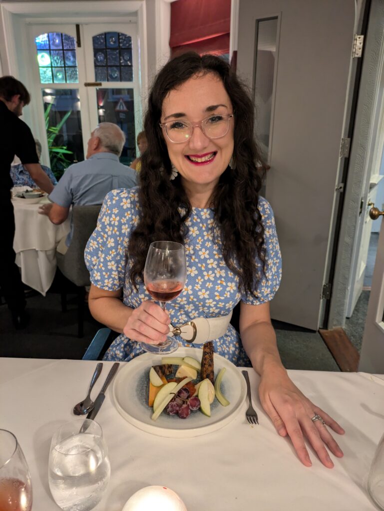 Georgina, a white brunette female who uses a wheelchair, raises a rose wine glass to the camera and smiles