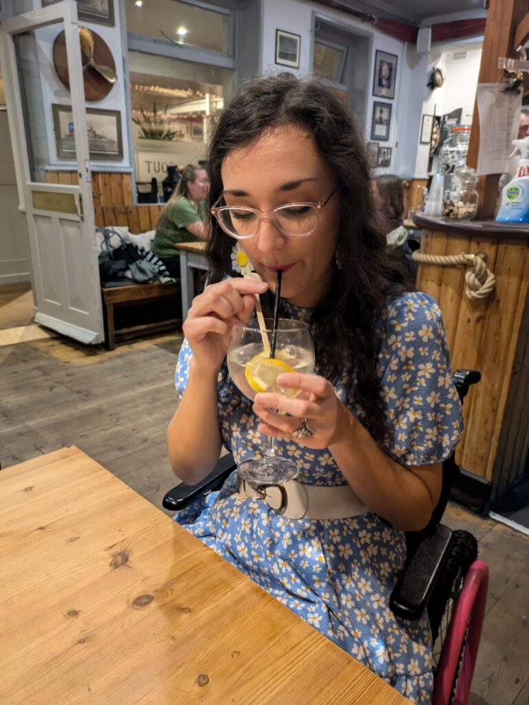 Georgina, a white brunette female who uses a wheelchair, sips a gin drink