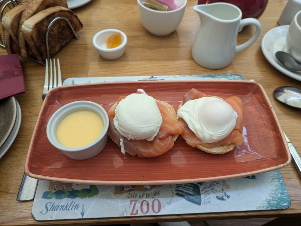 two poached eggs with salmon on English muffins, alongside a pot of Hollandaise sauce