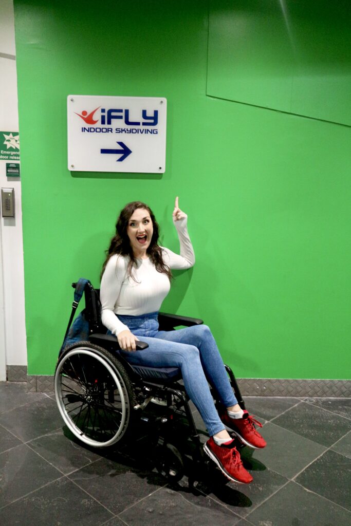 Georgina, a disabled brunette female in a casual outfit of red trainers, cream long-sleeved top and blue jeggings. She is smiling in front of a sign that reads iFly indoor sky-diving, pointing at the sign whilst sat in her manual wheelchair.