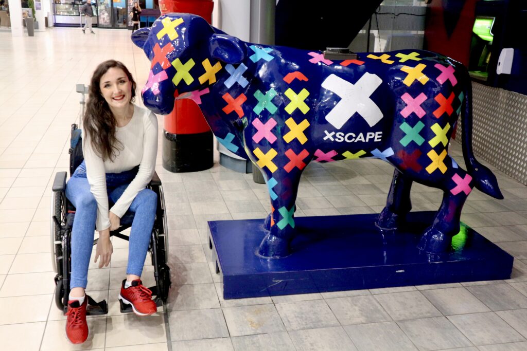 Georgina, a disabled brunette female in a casual outfit of red trainers, cream long-sleeved top and blue jeggings. She is smiling and leaning forward out of her manual wheelchair. She is next to a painted cow statue with the text Xscape on.