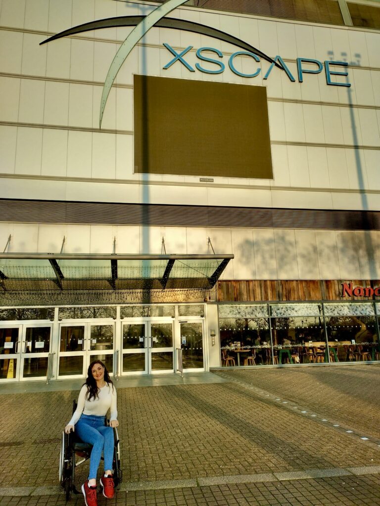 Georgina, a disabled brunette female in a casual outfit of red trainers, cream long-sleeved top and blue jeggings. She is smiling in front of the Xscape shopping centre entrance, sat in her manual wheelchair.