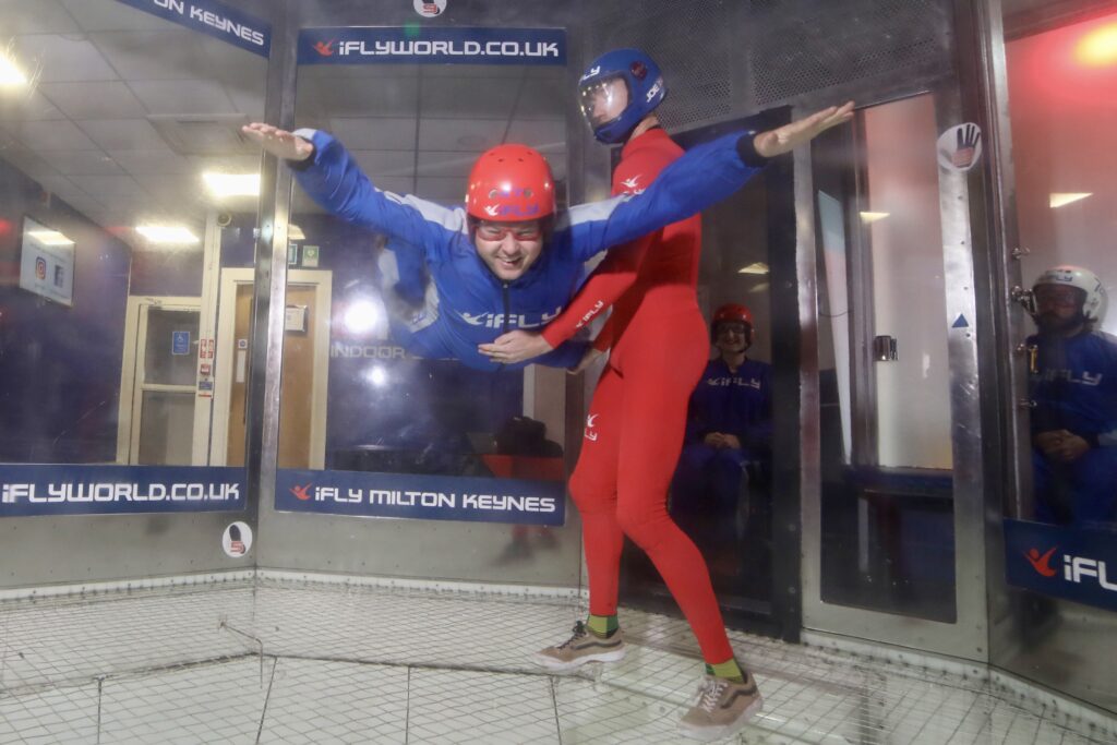 Richard, a brunette male in a blue and red flying suit. He is flying in an iFly indoor sky-diving chamber