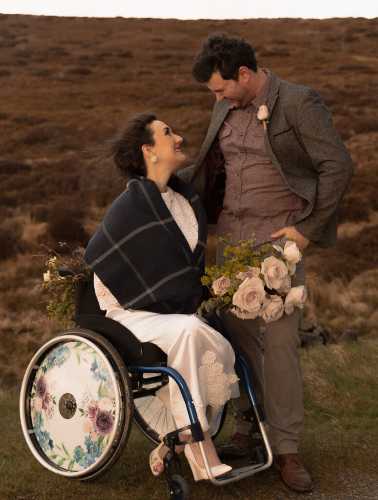 Georgina and Richard are modelling as a faux bride and groom. She is wearing a white dress and has floral wreath spoke guards on her wheelchair.