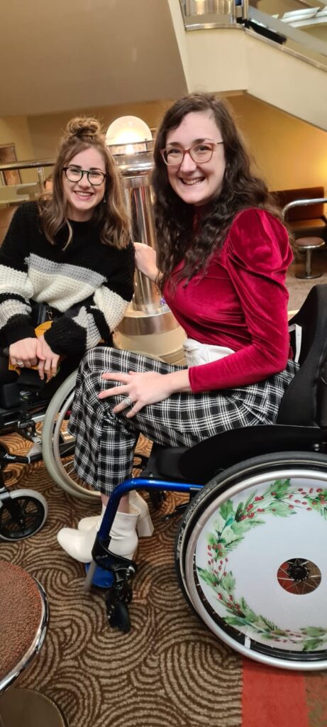 An image of two brunette young females. Both are smiling and Georgina sits in a manual wheelchair with spoke covers in a green wreath pattern.