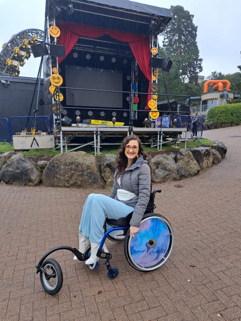 Georgina is a white brunette and is sat in a manual wheelchair. She has blue marble spoke guards on her manual wheelchair wheels.