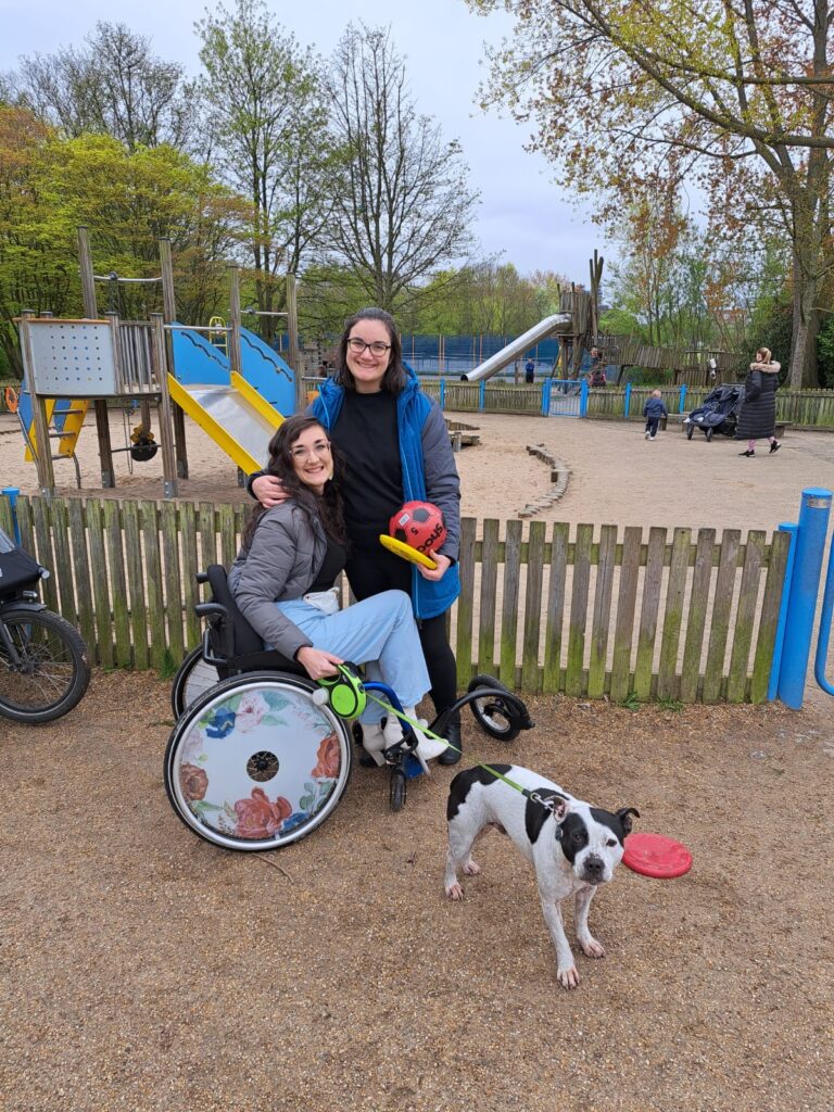 An image of two brunette females, one is a wheelchair user with colourful spoke guards on her chair. There is a Staffy dog.