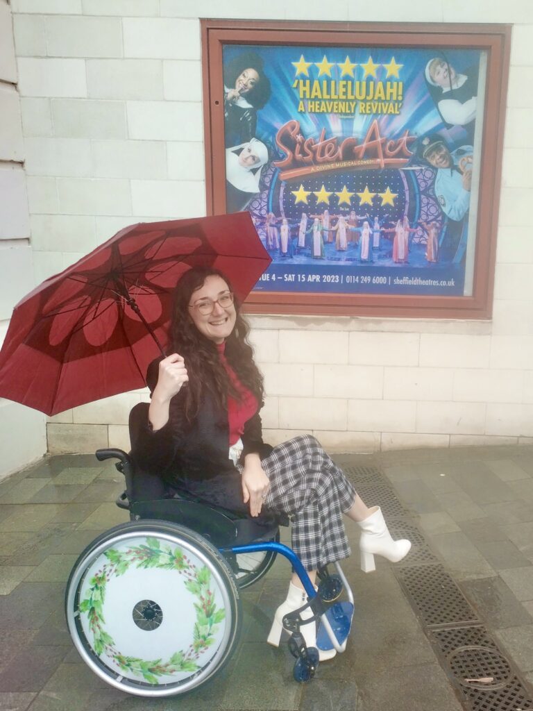 Georgina is a white brunette with a red umbrella. She has a festive design on her wheelchair spoke guards.