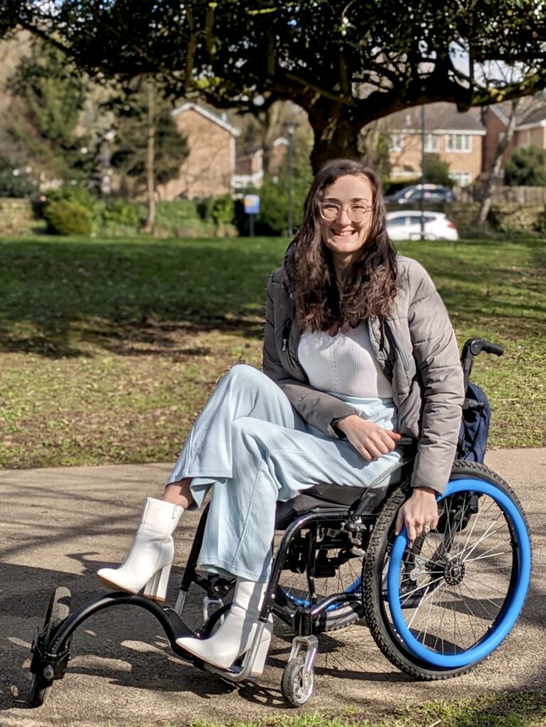 Georgina (a white brunette female) is outside with blue push rim covers on her wheelchair