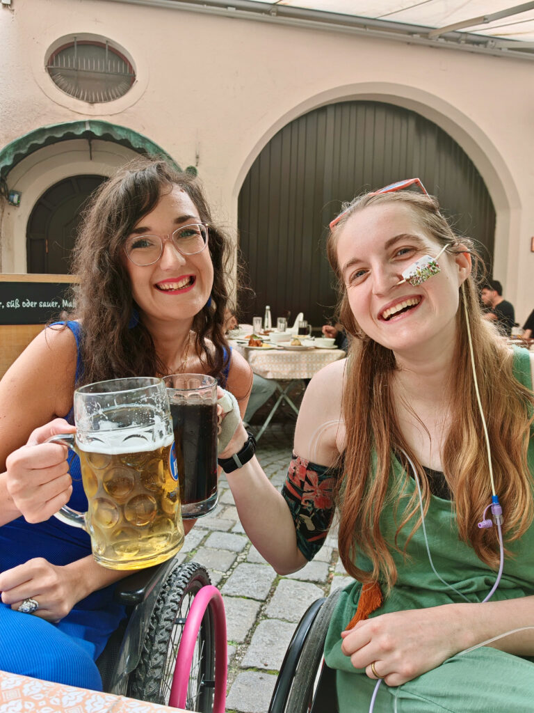 Mary and Georgina (two, white female manual wheelchair users) are doing a "cheers" with their steins