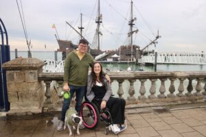 Georgina and Richard are smiling with a stunning Spanish Galleon ship behind them. They are by the sea and their staffy dog Milo is in front of them, on the lead.